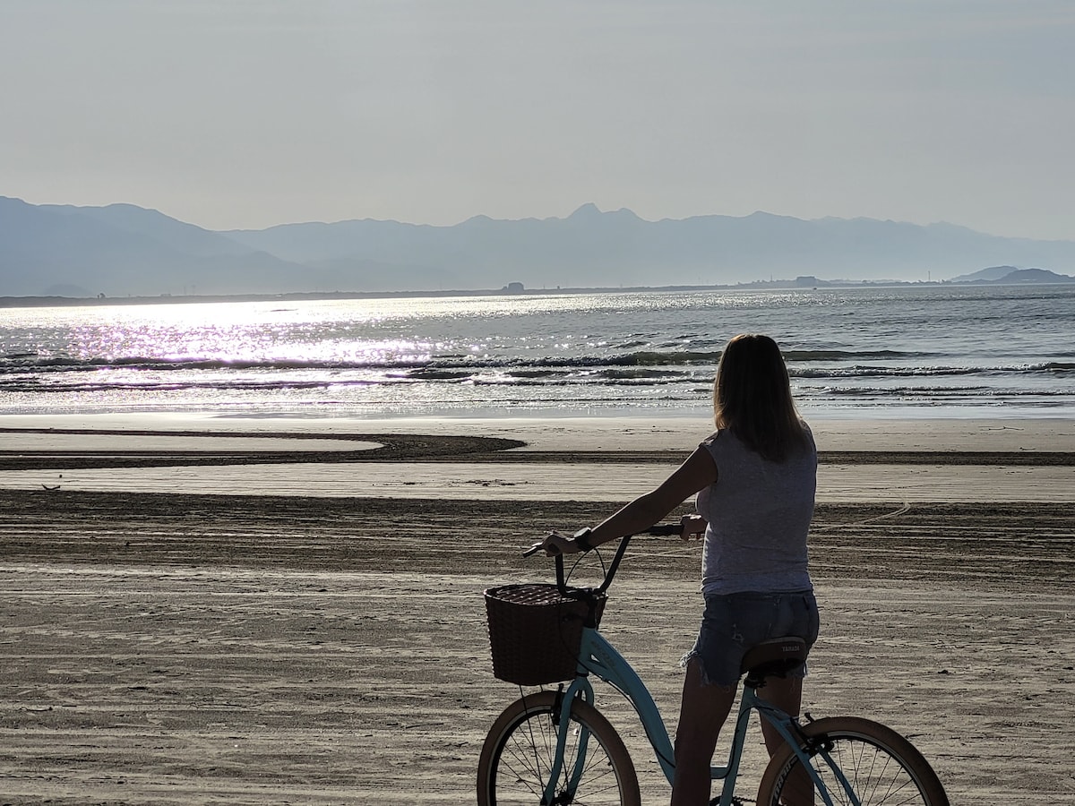 A cyclist can be seen on a sandy beach overlooking glistening waves. The distant mountains create a serene backdrop, reflecting the sunlight. Soft, rippling water extends into the horizon, where the sky meets the sea.