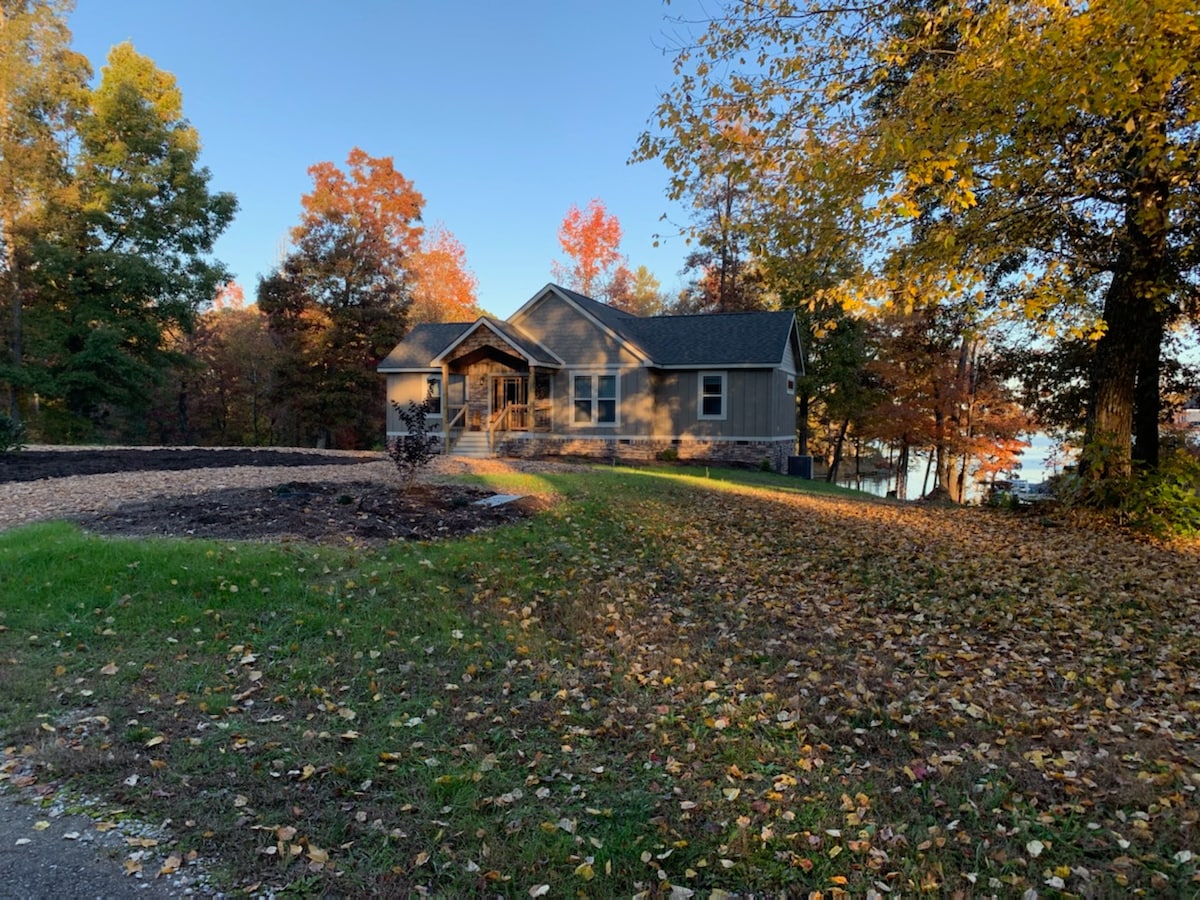 A custom vacation home is situated among vibrant autumn foliage. The exterior features a welcoming porch and a gently sloping yard, leading towards the serene waters of Watts Bar Lake, visible in the distance. The landscape is adorned with colorful leaves and well-maintained greenery.