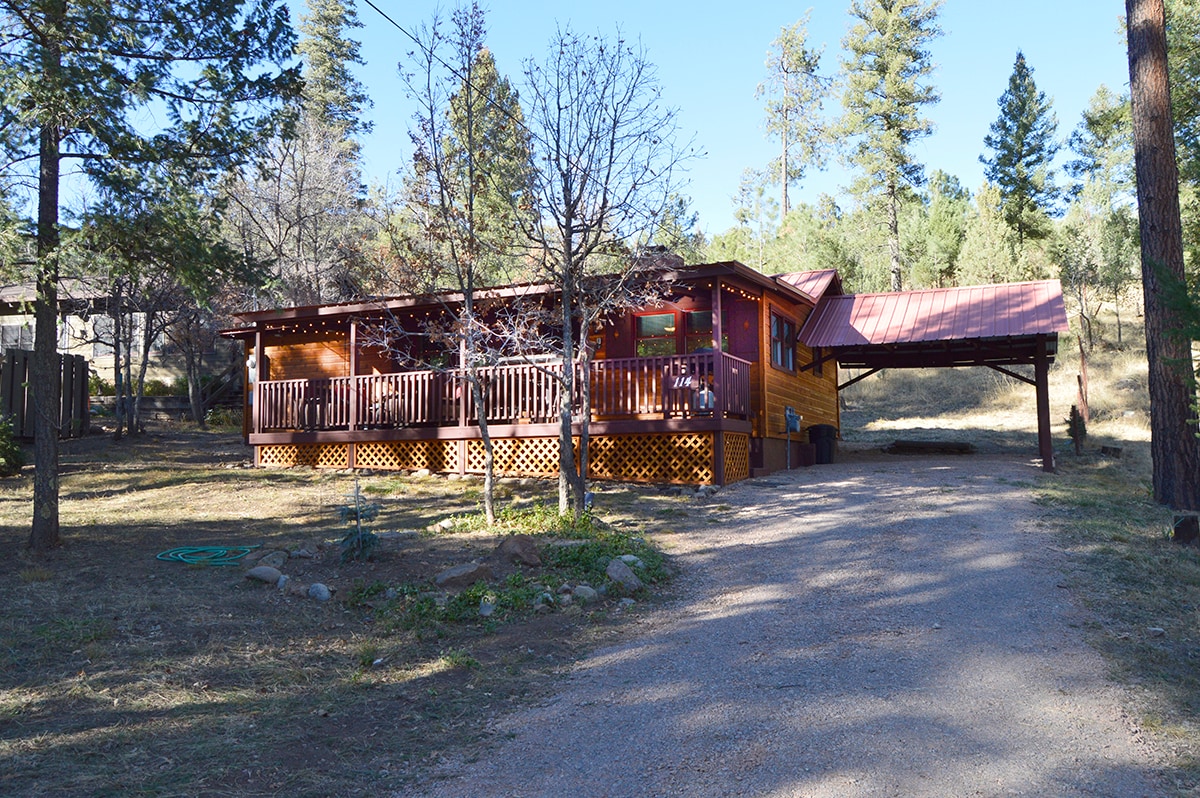 A cozy cabin exterior is viewed, featuring a wooden porch with a railing that spans the front. A carport is located on the right side, while a gravel driveway leads up to the cabin. Towering Ponderosa Pines surround the property, adding to the natural setting.
