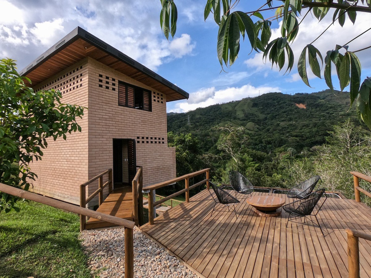 The exterior features a two-story cabaña constructed of light-colored brick, set amidst lush greenery. A wooden terrace is visible, furnished with stylish seating and a round table, offering views of the surrounding mountains and sky. Natural light brightens the structure, enhancing its connection with nature.