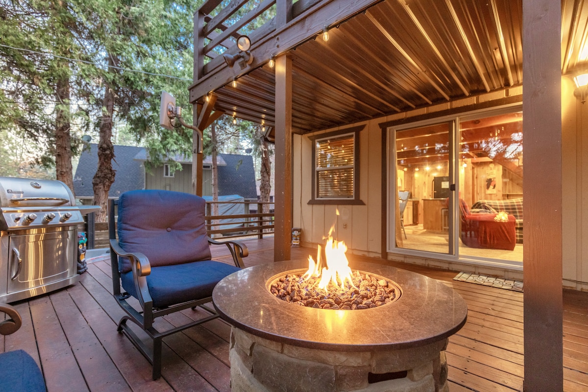 An outdoor deck features a stone fire pit with a flickering flame at its center, surrounded by blue seating. A gas grill is positioned nearby, and large windows provide a view of the cozy living space inside, framed by tall trees.