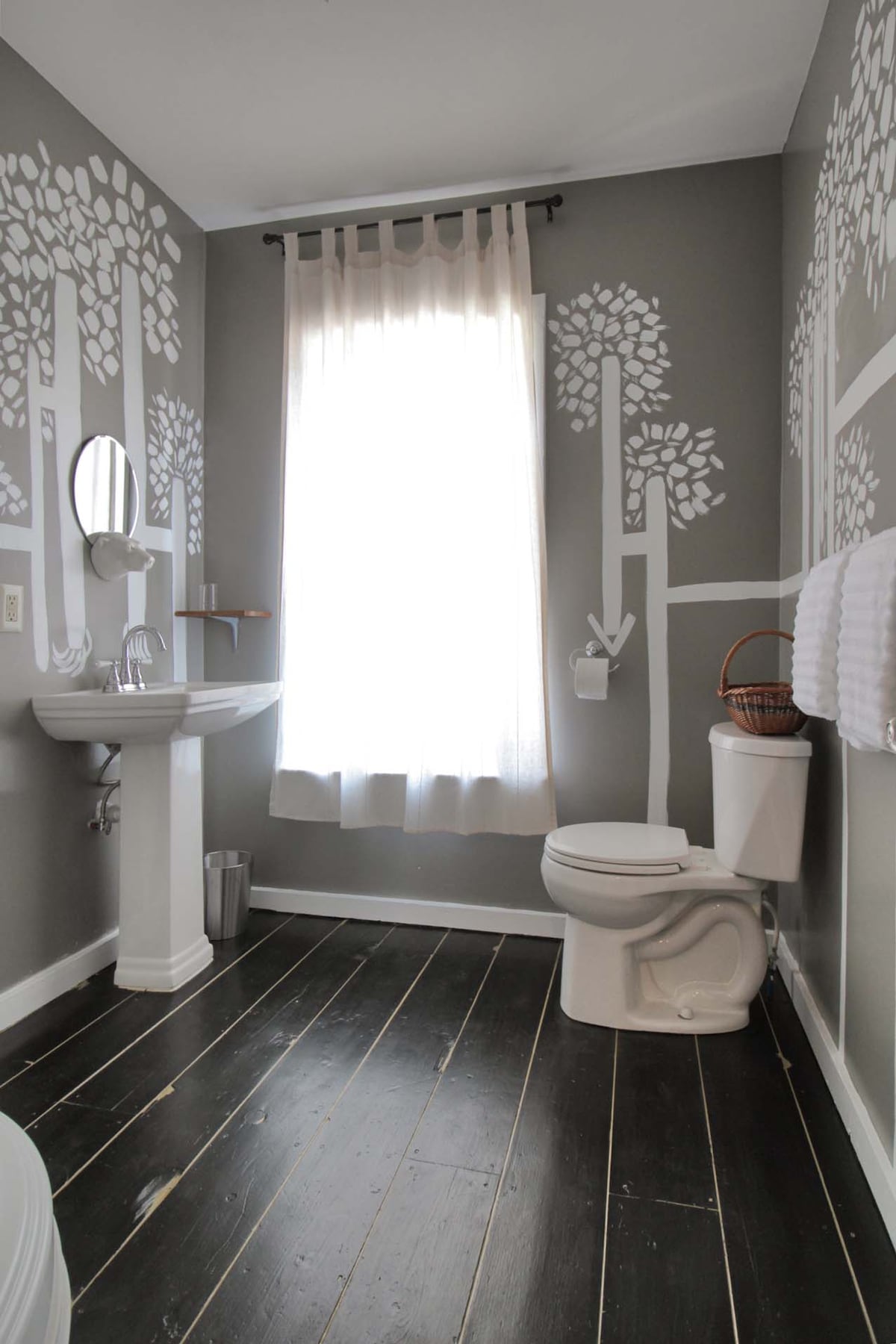 A well-appointed bathroom features tree-themed wall art against muted gray tones. A freestanding sink is accompanied by a round mirror, and a white shower curtain provides privacy. Natural light filters through a sheer curtain, illuminating dark wooden flooring and adding a touch of warmth.