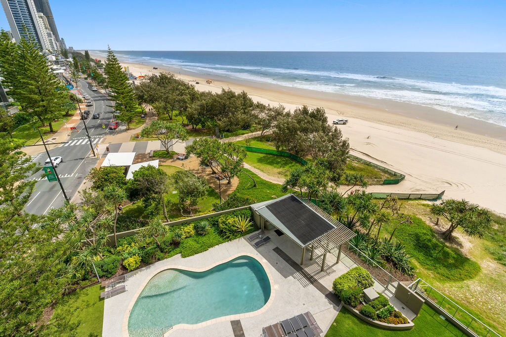 An aerial view showcases a landscaped area featuring a heated pool beside a BBQ facility, with loungers arranged around it. The beach stretches into the distance, bordered by gently waving trees and a sandy shore. Coastal activity can be seen along the beach and water.
