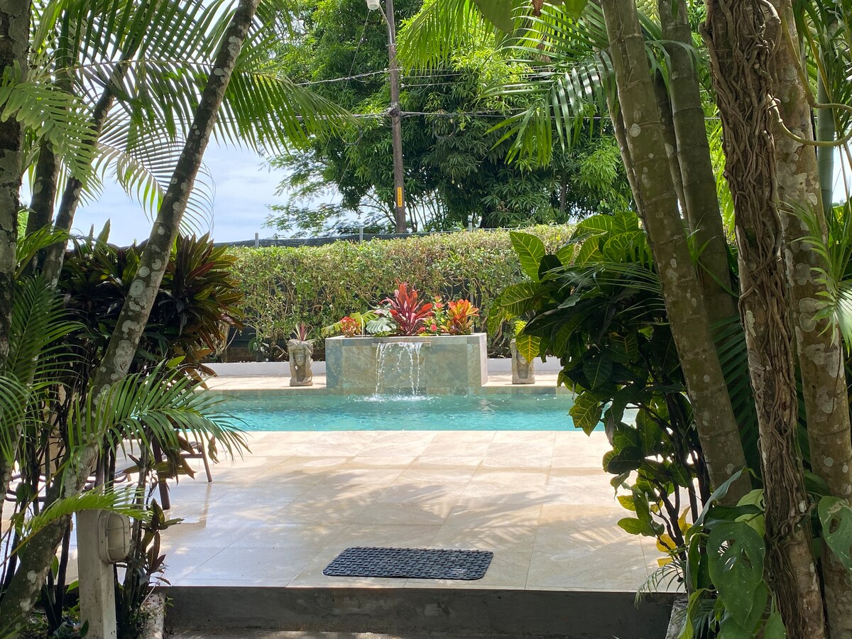 A serene view of the pool area framed by lush tropical foliage. Water flows gently from a decorative feature into the clear water, which reflects the surrounding greenery. The pool deck is spacious and inviting, offering a peaceful atmosphere.