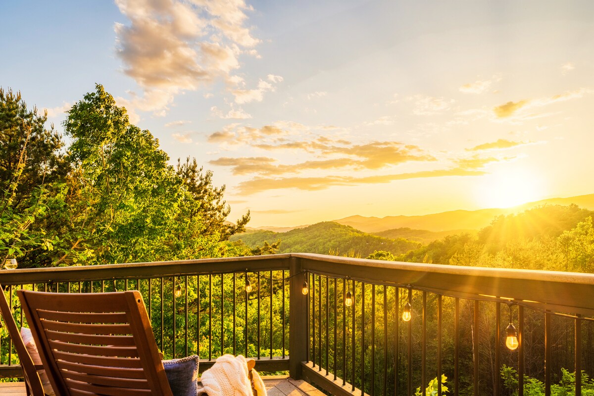 A wooden deck showcases a view of rolling mountains at sunset, with golden light streaming through clouds. The railing is adorned with light bulbs, and a cozy chair is positioned for relaxation, inviting guests to appreciate the surrounding nature.