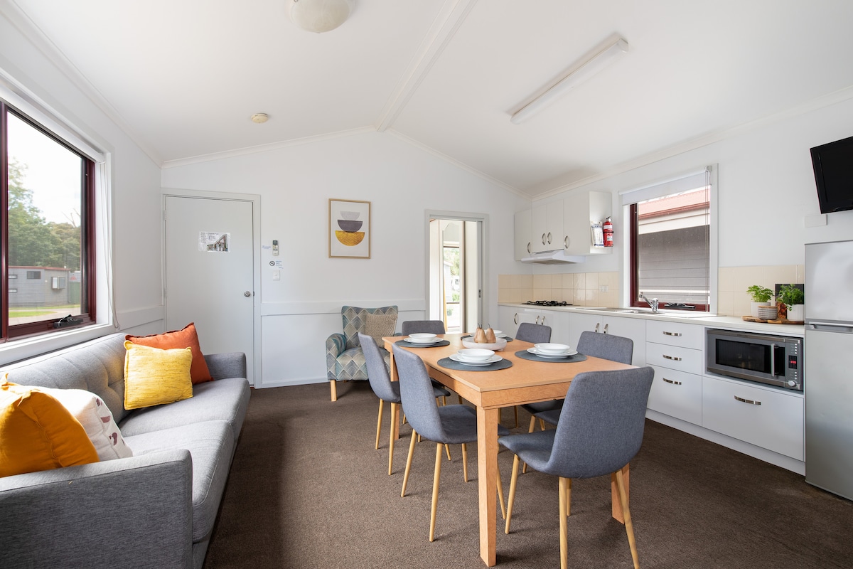 A bright and airy living space is presented, featuring a comfortable gray sofa with accent pillows. A dining table with six chairs is arranged nearby. A kitchenette with white cabinetry and appliances is visible in the background, illuminated by natural light from surrounding windows.
