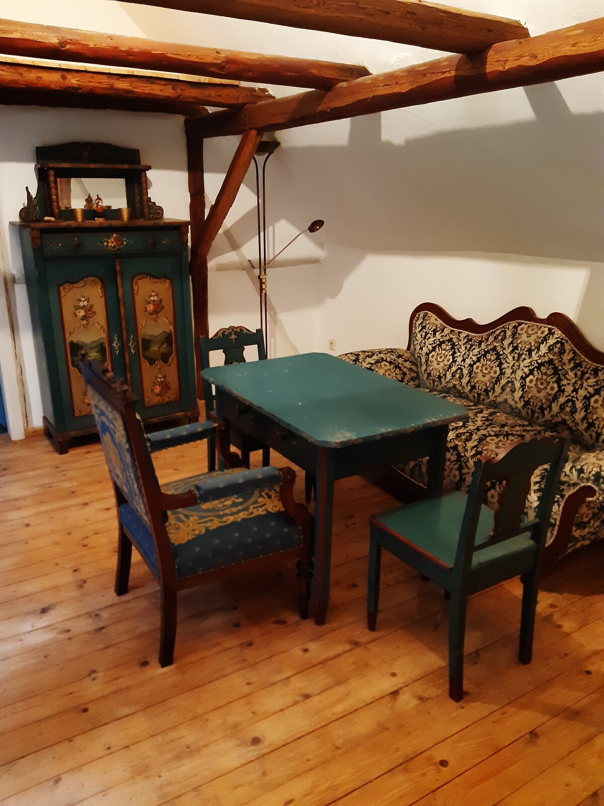 A dining area is presented with a rectangular table surrounded by four chairs, featuring ornate designs. A patterned sofa is placed against the wall. The rustic wooden beams and warm wooden flooring enhance the cozy atmosphere of the space.