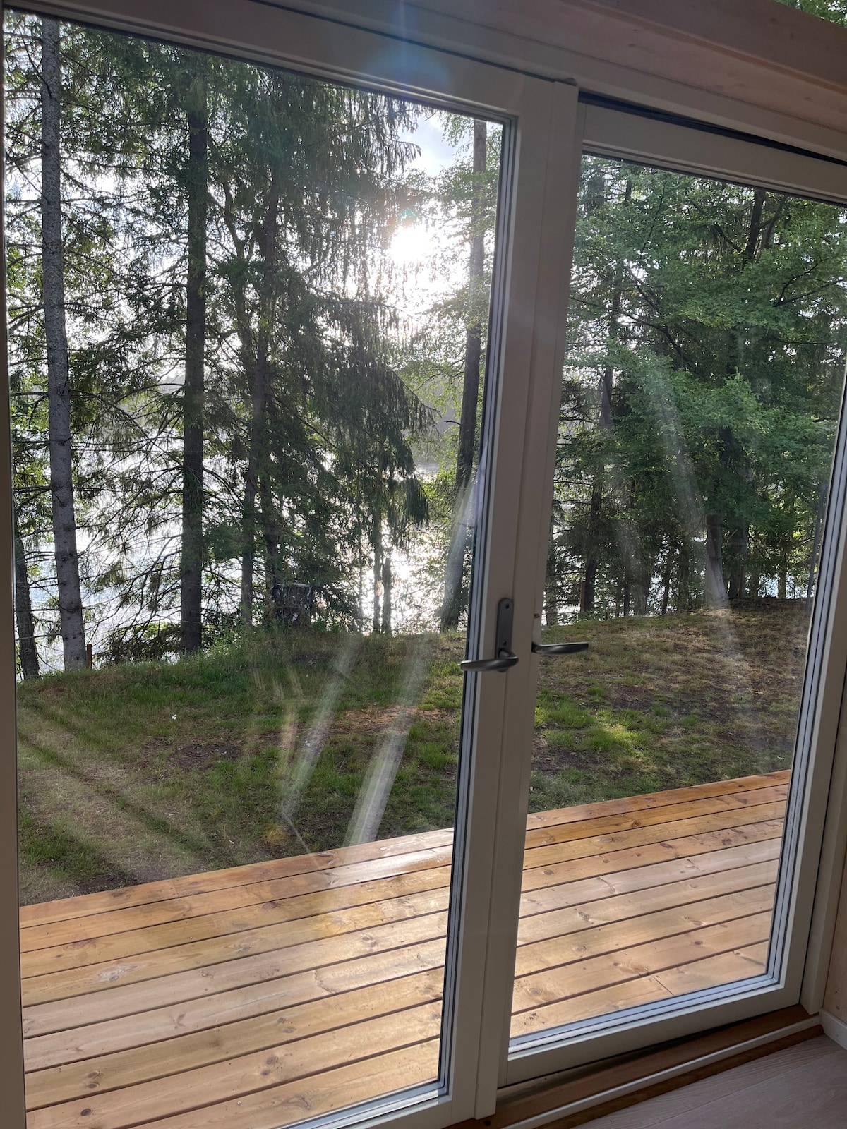 A double glass door opens to a wooden deck, framed by trees. Natural light filters through the glass, casting reflections on the floor. Beyond, the serene surface of the lake glistens in the distance, surrounded by lush greenery.