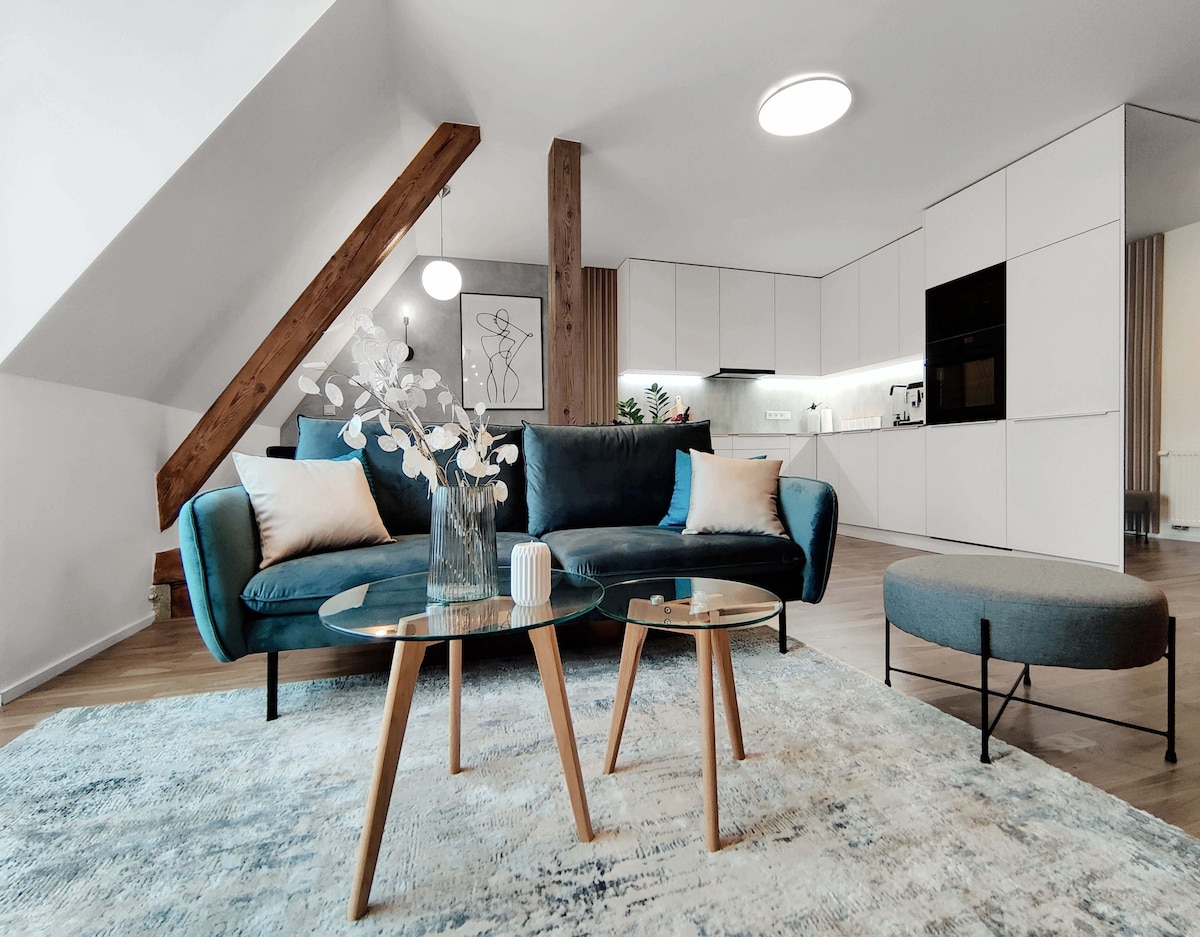 A modern living space features a teal sofa adorned with neutral cushions, positioned across from two circular glass tables. A round ottoman complements the decor. Soft lighting is provided by a ceiling fixture, while wooden beams add character to the room.