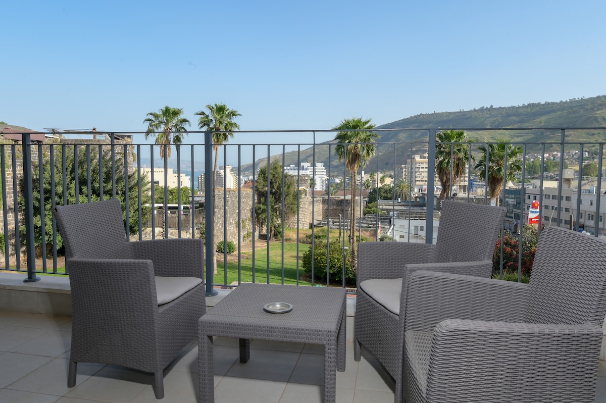 A terrace is furnished with two armchairs and a small table, surrounded by palm trees. The view includes green hills and a distant glimpse of the sea, providing a serene outdoor space for relaxation.
