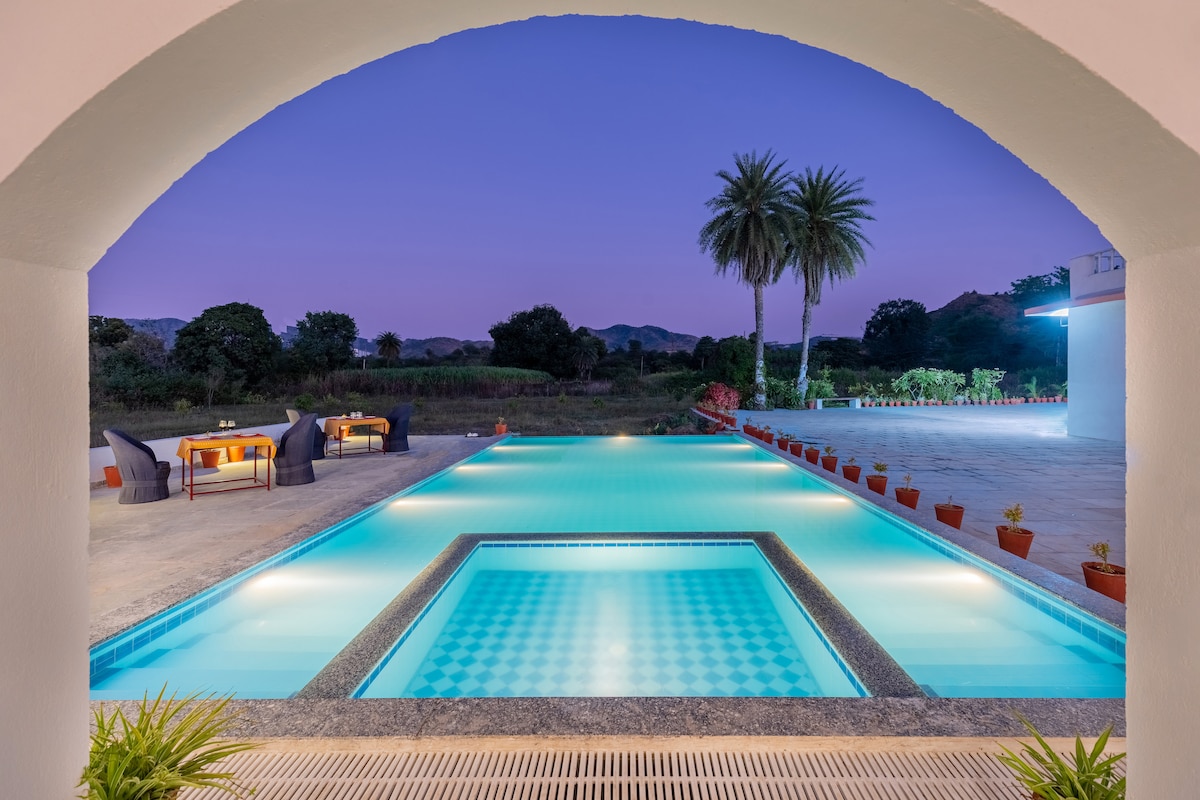 An inviting outdoor area features a tranquil pool with a geometric design, surrounded by lush greenery. Comfortable seating areas are visible in the background, complemented by potted plants. The scene is framed by an archway under a calming twilight sky.