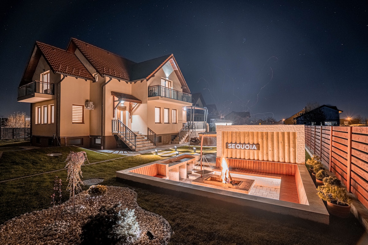 A modern villa is illuminated at night, featuring a private hot tub area and an individual fire pit surrounded by pavers. The spacious yard is landscaped with decorative rocks and plants, while the inviting exterior of the villa showcases large windows and multiple balconies.