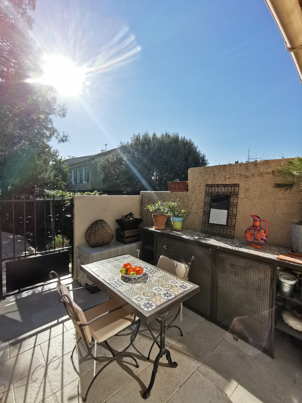 An outdoor dining area is showcased under a bright sun. A metal table with patterned tiles is surrounded by two chairs. Potted plants add greenery to the space, while a mirror reflects light from the warm sun. A stone wall provides privacy.