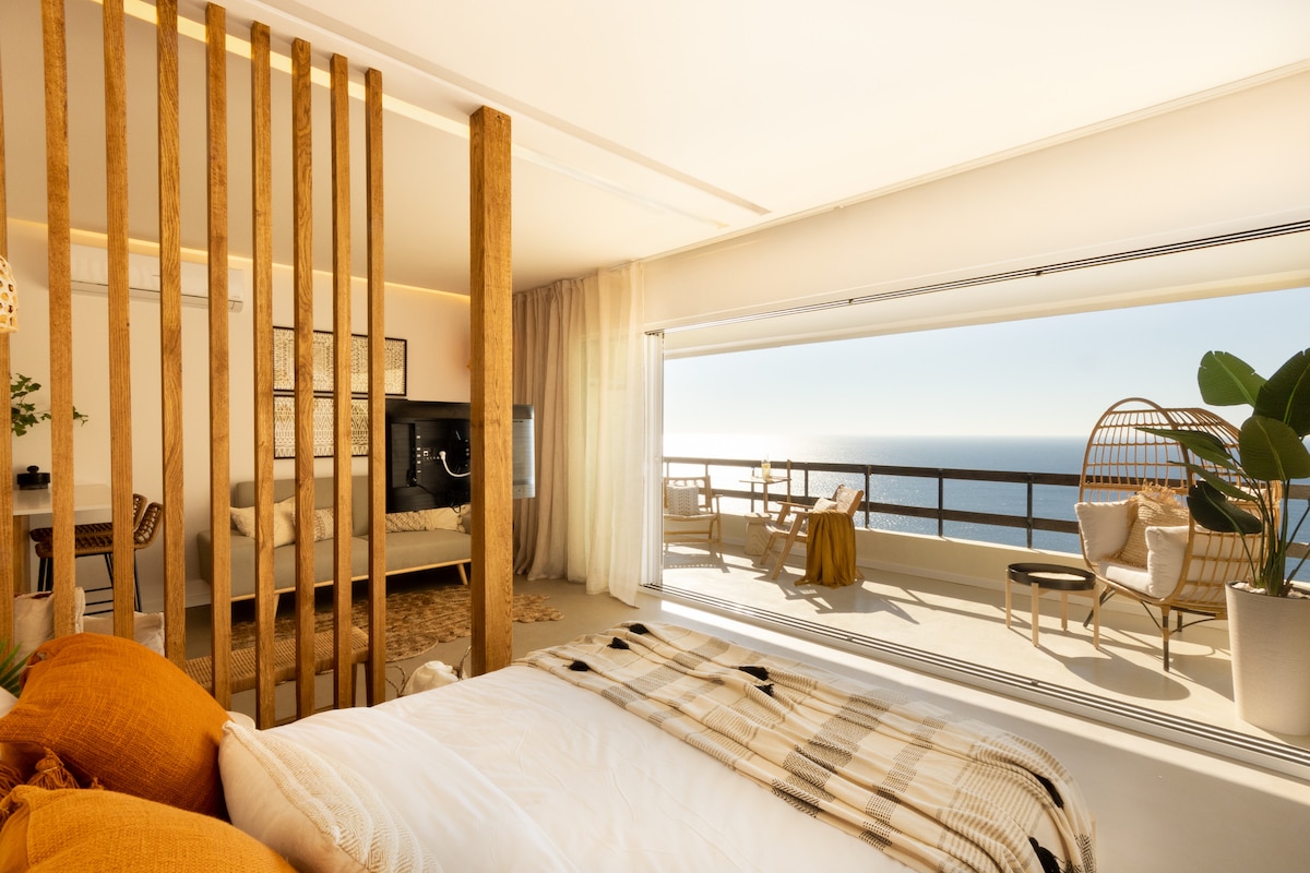A spacious studio is highlighted by large glass doors offering expansive views of the ocean. The sleeping area features a comfortable bed with neutral bedding, complemented by soft textures and natural light. A balcony is adorned with stylish outdoor seating, inviting relaxation.