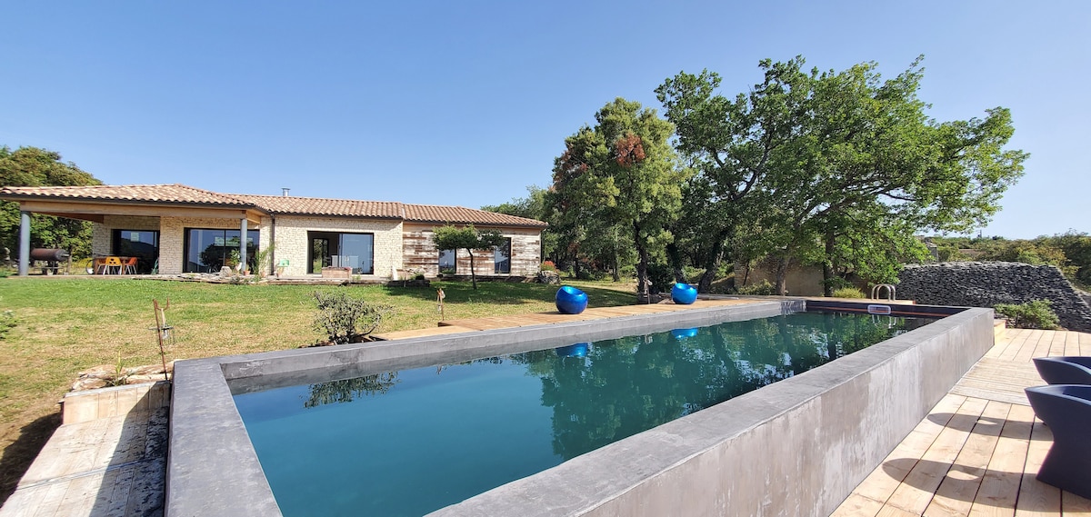 A long swimming pool is set against a backdrop of lush greenery and a single-story house. The pool features a sleek, modern design with water reflecting the clear blue sky. A few outdoor seating areas, including round chairs, are visible nearby.
