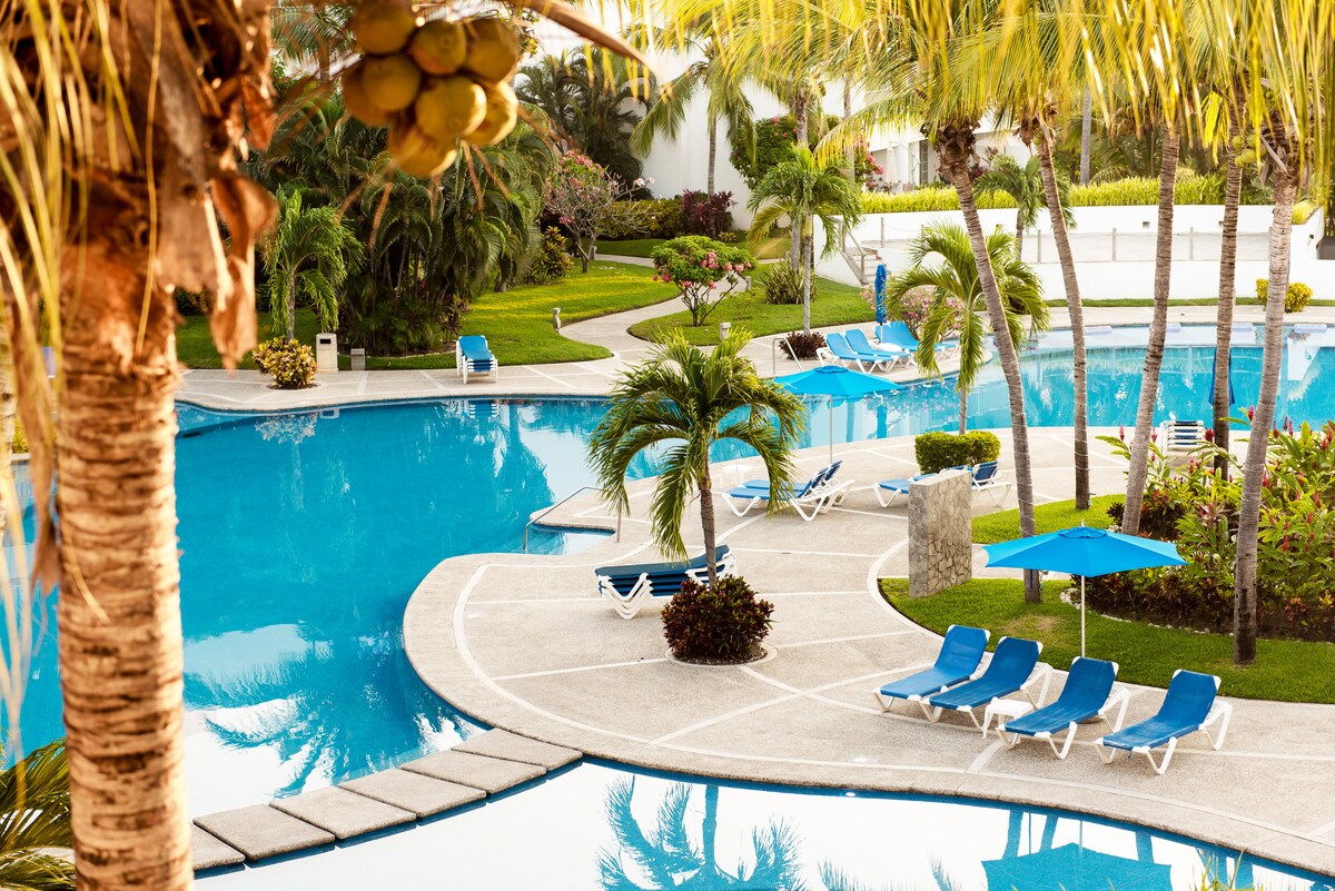 A tranquil scene is presented featuring two large swimming pools surrounded by vibrant greenery and palm trees. Lounge chairs are arranged along the pool edges, while shaded tables are positioned for relaxation. The lush landscaping enhances the serene environment.