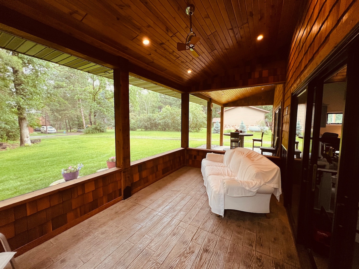 A covered porch area features warm wood paneling and a comfortable white sofa. Large windows provide views of the lush green yard, allowing natural light to fill the space. Potted plants are positioned along the porch, enhancing the serene atmosphere.