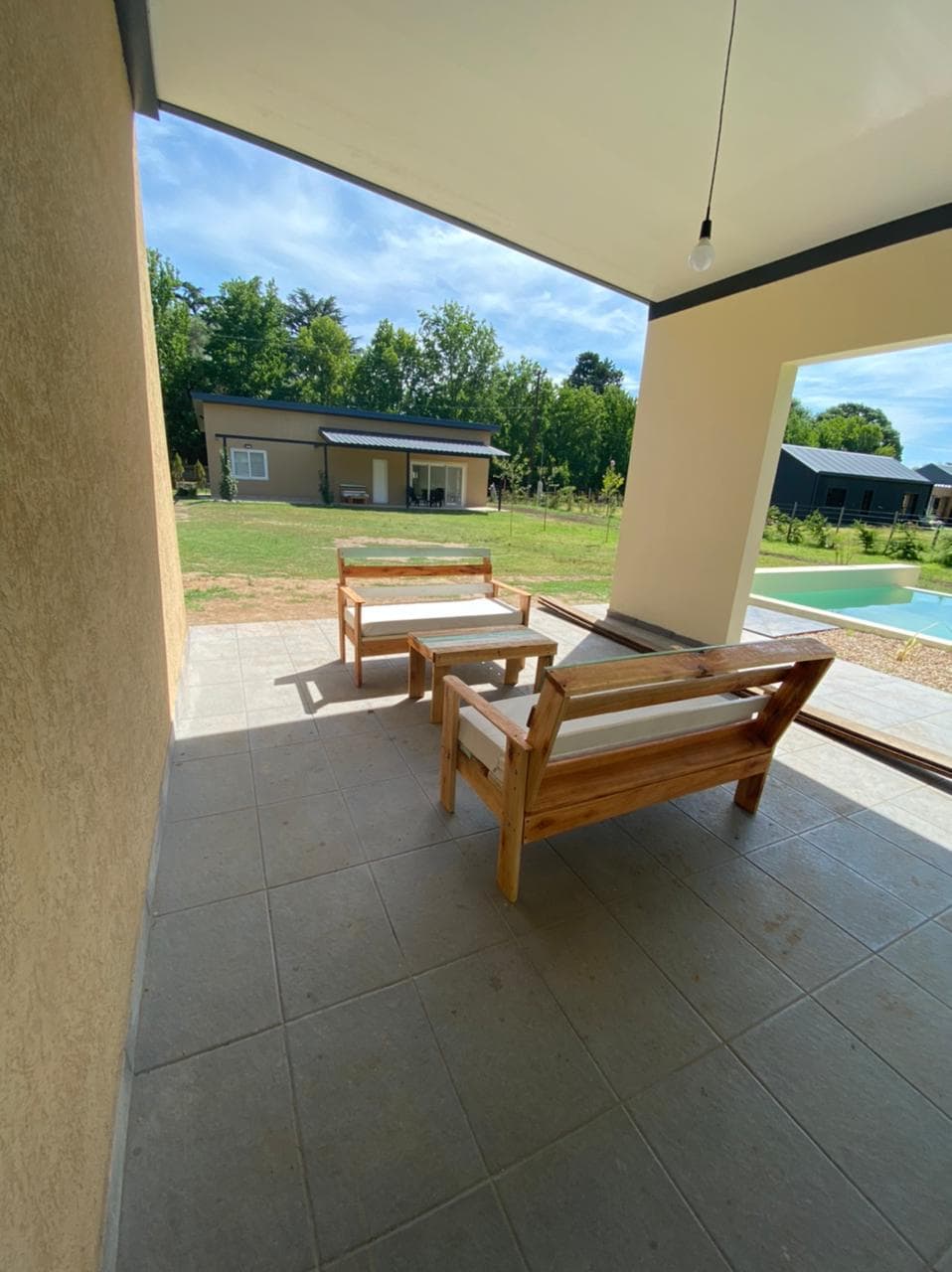 A covered outdoor seating area is shown, featuring two wooden lounge chairs with light cushions. The space is surrounded by greenery, with a view of nearby cabins and a swimming pool in the background. Natural light illuminates the area, enhancing the relaxing atmosphere.