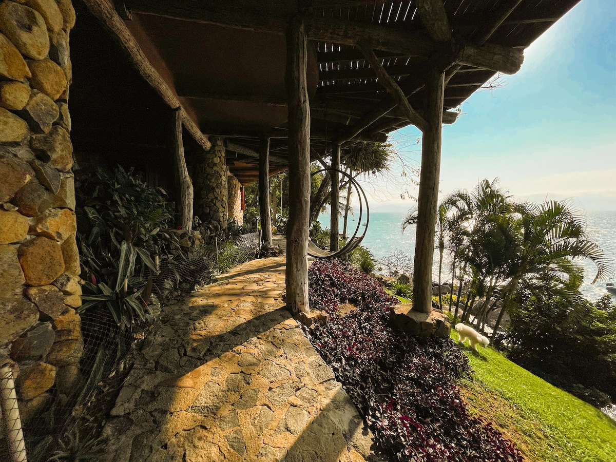 A stone path winds alongside lush greenery, leading to the stunning ocean view. The structure features rustic wooden beams, seamlessly integrating natural elements. The vibrant foliage and tropical plants create a serene atmosphere, inviting guests to enjoy the tranquil coastal scenery.