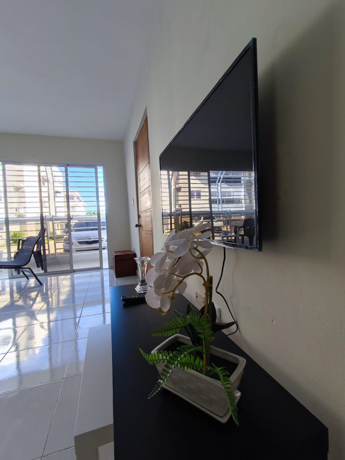 A modern living area features a flat-screen TV mounted on the wall above a sleek black console table. A small potted plant adds a touch of greenery, while natural light streams through the large windows, illuminating the open space.