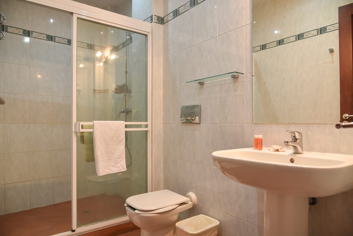 A clean and functional bathroom features a glass shower enclosure, a white toilet, and a modern sink. Light tiles create a bright atmosphere, while a towel hangs conveniently on the shower door. A small container for toiletries is placed near the sink.