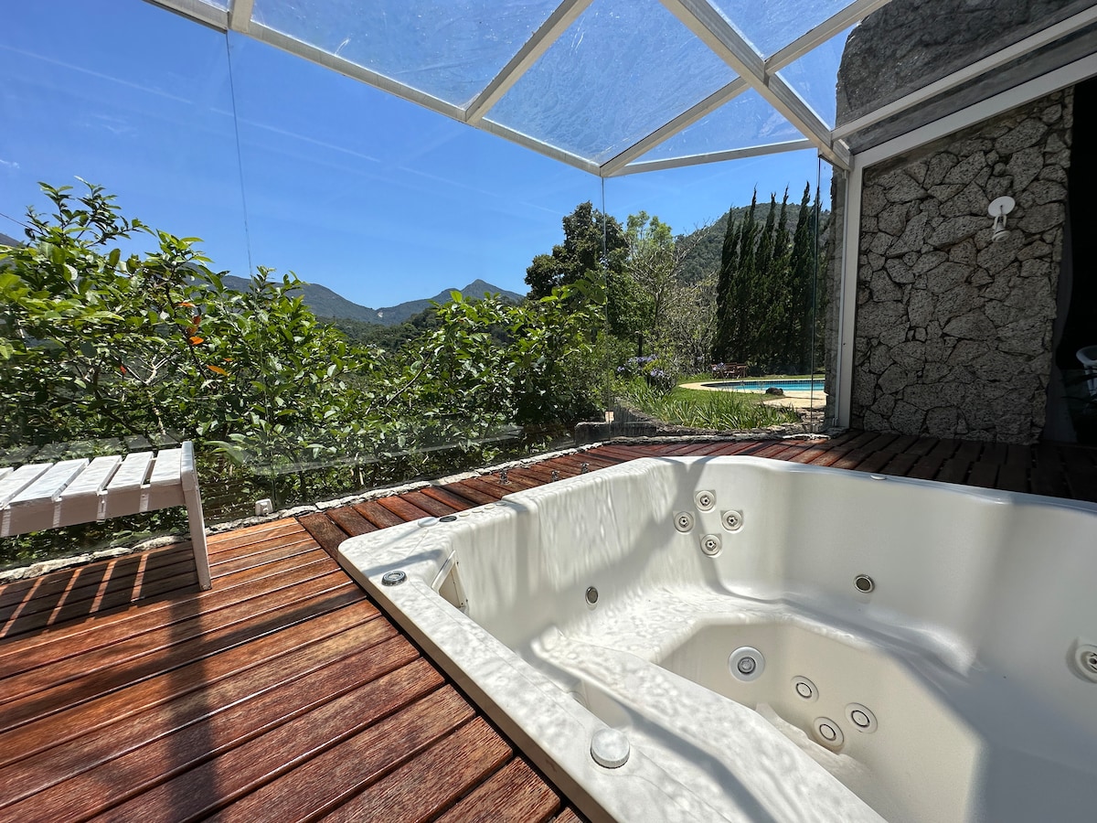 A spacious hot tub is situated on a wooden deck, surrounded by greenery and offering views of the mountains and trees. The area is illuminated by natural light entering through the clear roof, enhancing the sense of relaxation.
