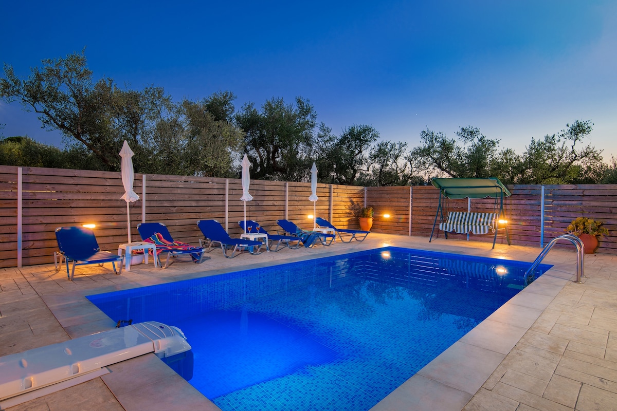 A private outdoor pool is surrounded by comfortable lounge chairs beneath soft lighting. The pool features a vibrant blue hue, while a shaded swing offers a relaxing seating option. Lush greenery is visible in the background, enhancing the tranquil setting.