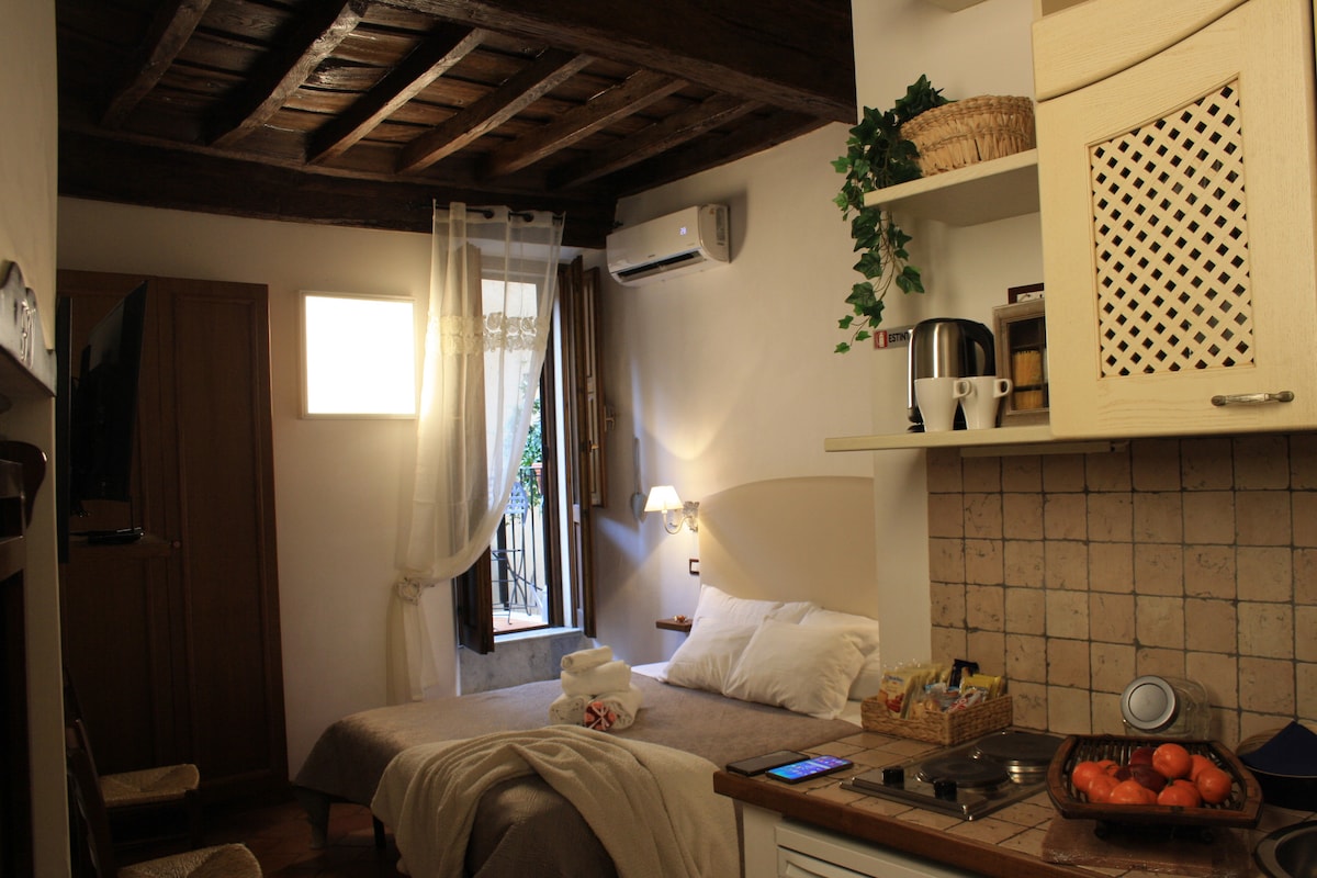 A cozy loft space features wooden ceilings and soft lighting. A bed with fresh linens is positioned next to a window draped with sheer curtains. A compact kitchen area includes a countertop, kettle, and a basket. Decorative plants are placed on shelves, enhancing the ambiance.