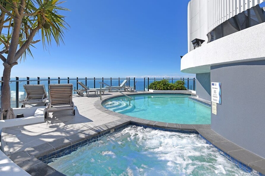 An outdoor rooftop pool area is depicted, featuring a shimmering pool with a spa section. Comfortable lounge chairs are arranged around the pool, complemented by a clear blue sky and ocean views. A palm tree adds a tropical touch to the relaxing ambiance.