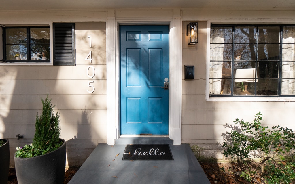 The entrance features a vibrant blue door framed by large windows on either side. A welcome mat with the word 'hello' is positioned at the base of the door, surrounded by potted greenery and a simple stone pathway leading to the door.