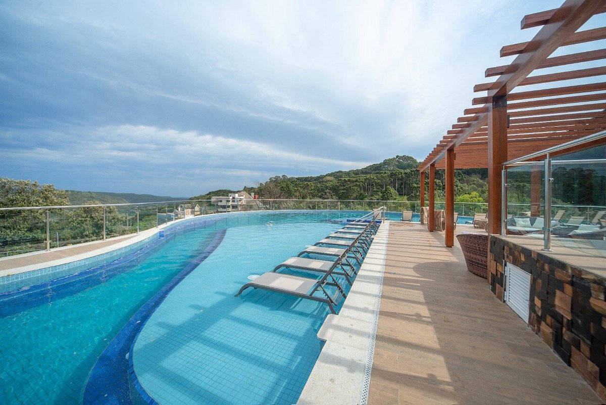 A modern infinity pool is framed by wooden decking and overlooks a scenic landscape. Sun loungers are positioned beside the pool, inviting relaxation. Large glass panels provide unobstructed views, enhancing the serene atmosphere of the space.
