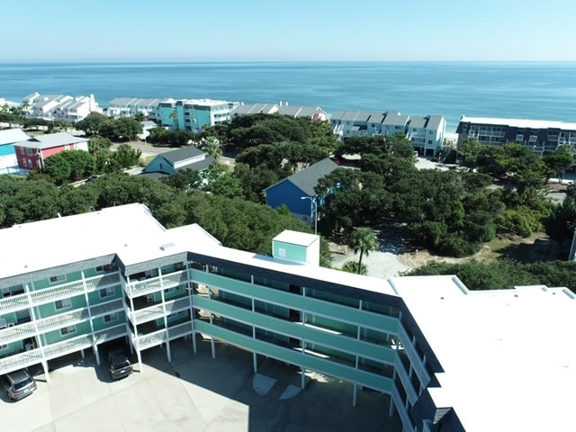 An aerial view showcases the modern beach condo amidst lush greenery and facing the ocean. The building features multiple levels with balconies overlooking the scenic coastline. The surrounding area includes colorful beach houses and well-maintained landscaping, emphasizing the tranquil beachside setting.