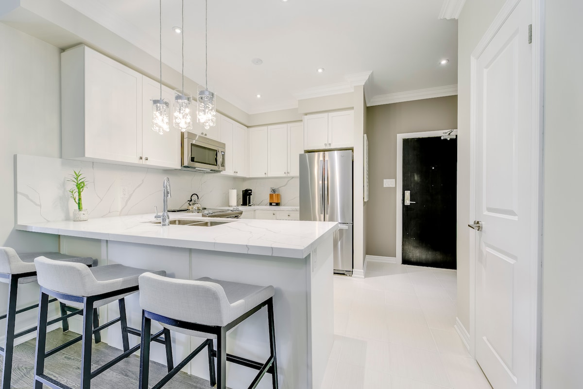 A modern kitchen features sleek white cabinetry and a large island with seating for four. Stainless steel appliances are visible, including a refrigerator and microwave. Bright lighting fixtures hang above the island, and a small green plant adds a touch of freshness to the countertop.