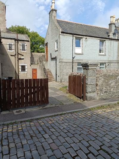 A quiet cul de sac is depicted, featuring a stone-paved street with a mix of building styles. The light blue two-story building stands beside a wooden fence, with wooden stairs leading to an entrance. Nearby, a red door is visible on another building.