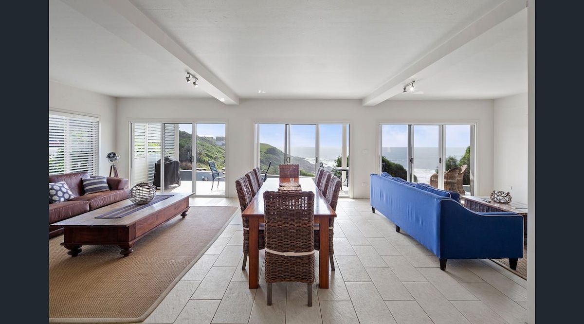 A spacious open living area features large glass doors that provide views of the coastline. Dark wood and woven furniture offer seating options around a central dining table. Natural light fills the room and highlights the tiled flooring, creating a bright and airy atmosphere.