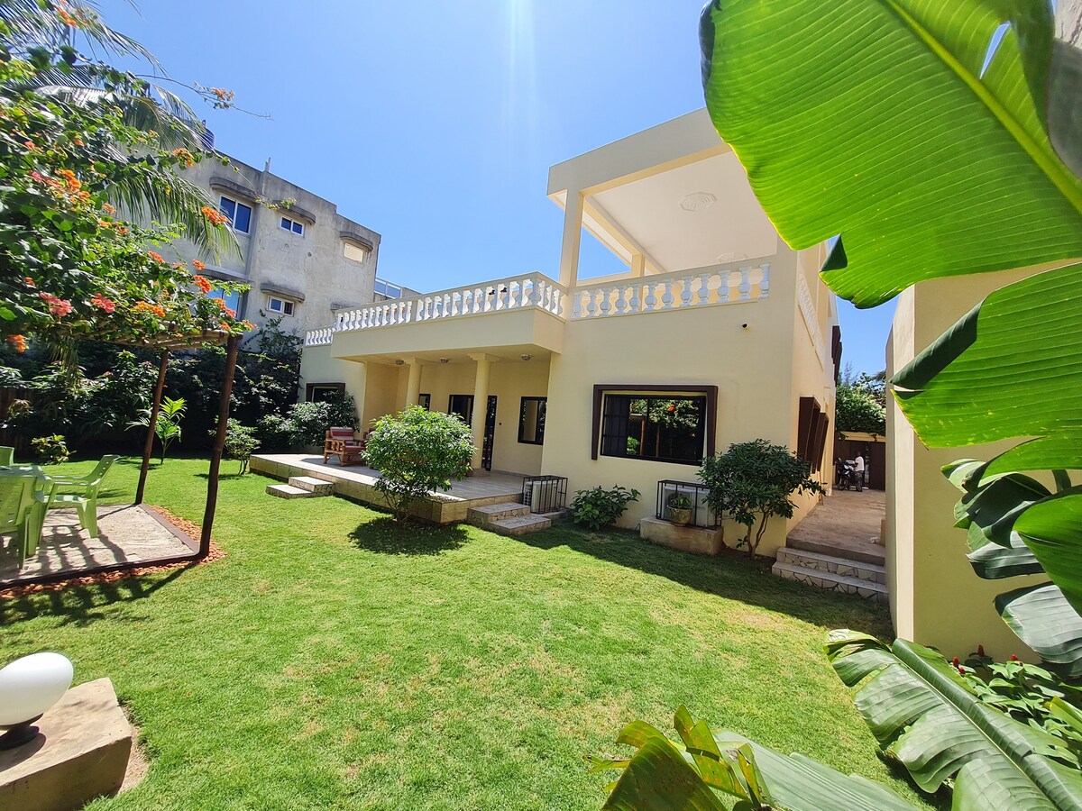 The exterior of the villa is visible, featuring a large garden with well-maintained green grass. The two-story structure includes a spacious balcony with railing and multiple large windows. Surrounding foliage adds to the serene setting.