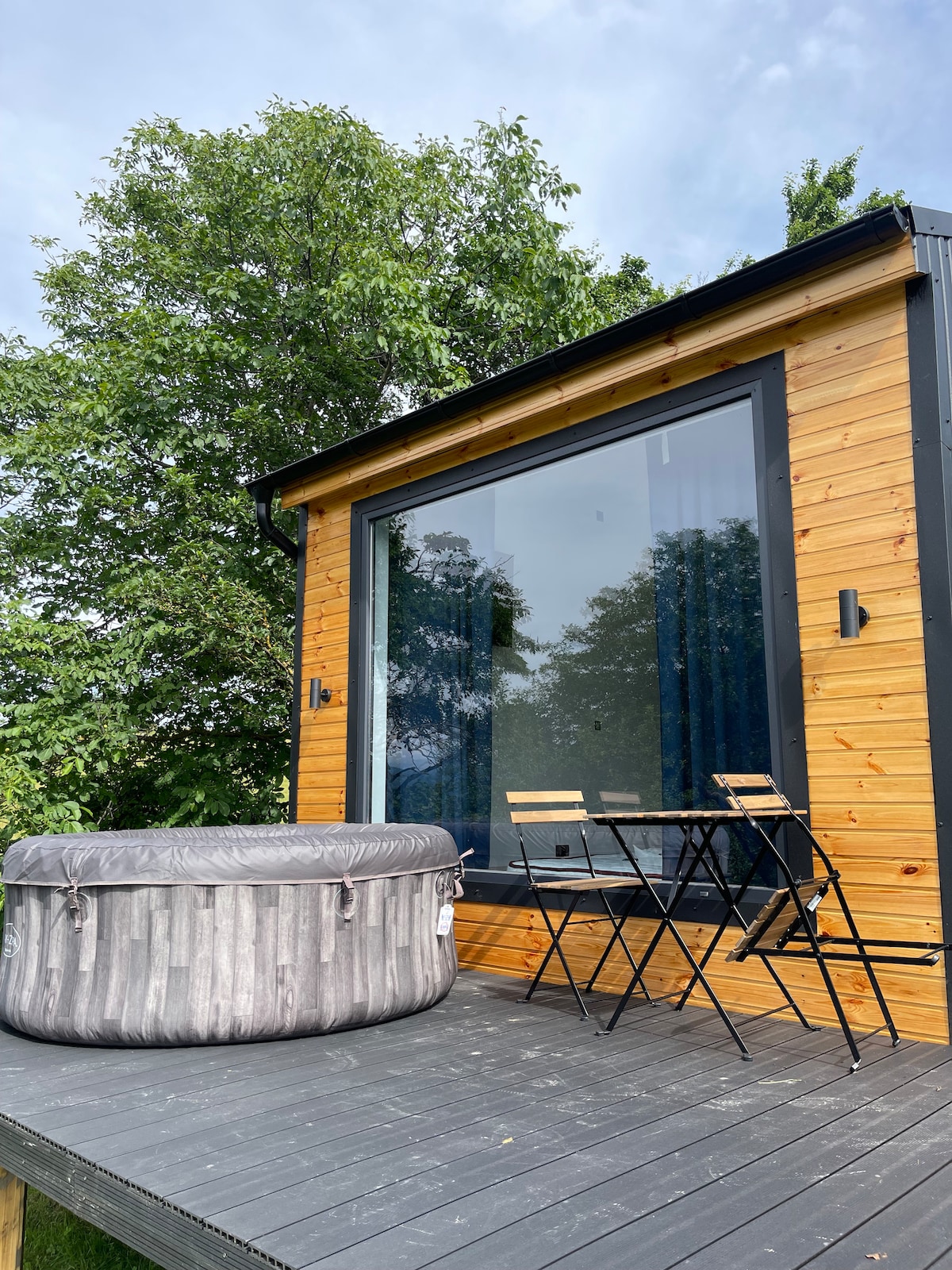 A wooden deck features a round, inflatable hot tub and a small, round table with two chairs. Large glass doors provide a view into the tiny chalet, framed by lush greenery from nearby trees.