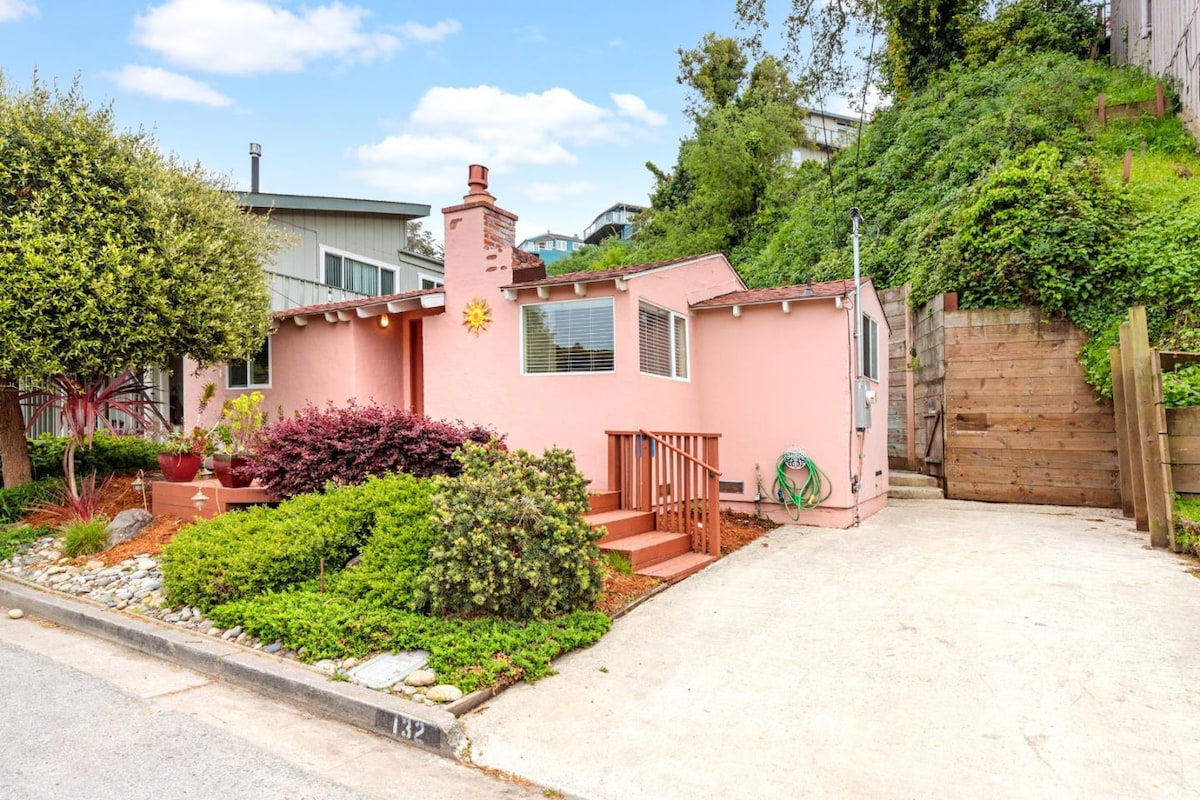 A charming, single-story home features a pastel pink exterior, adorned with a sun motif. Lush greenery surrounds the property, complemented by a gravel path. A private driveway offers parking space, while steps lead to the front entrance, enhancing its welcoming appearance.