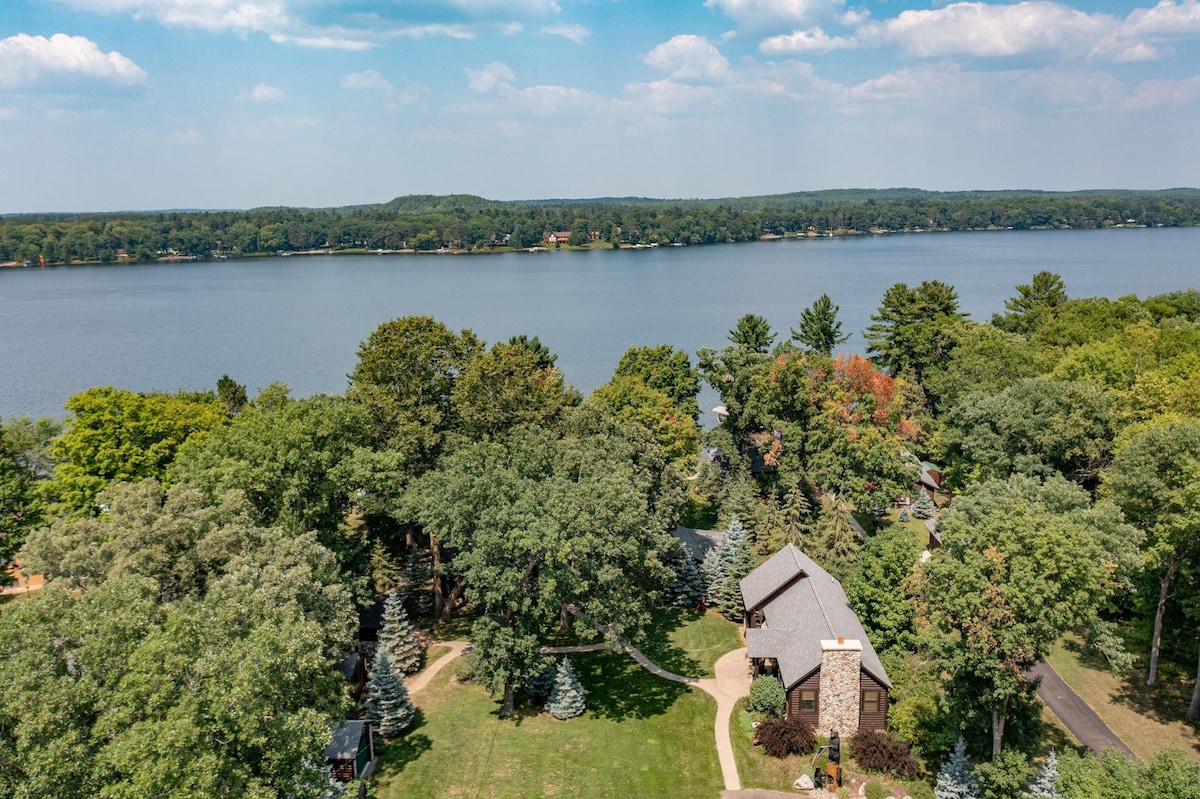An aerial view captures the cabin nestled among trees with vibrant greenery, facing a calm lake under a blue sky dotted with clouds. The property features a winding driveway leading to the entrance, with additional cabins visible in the surrounding area.