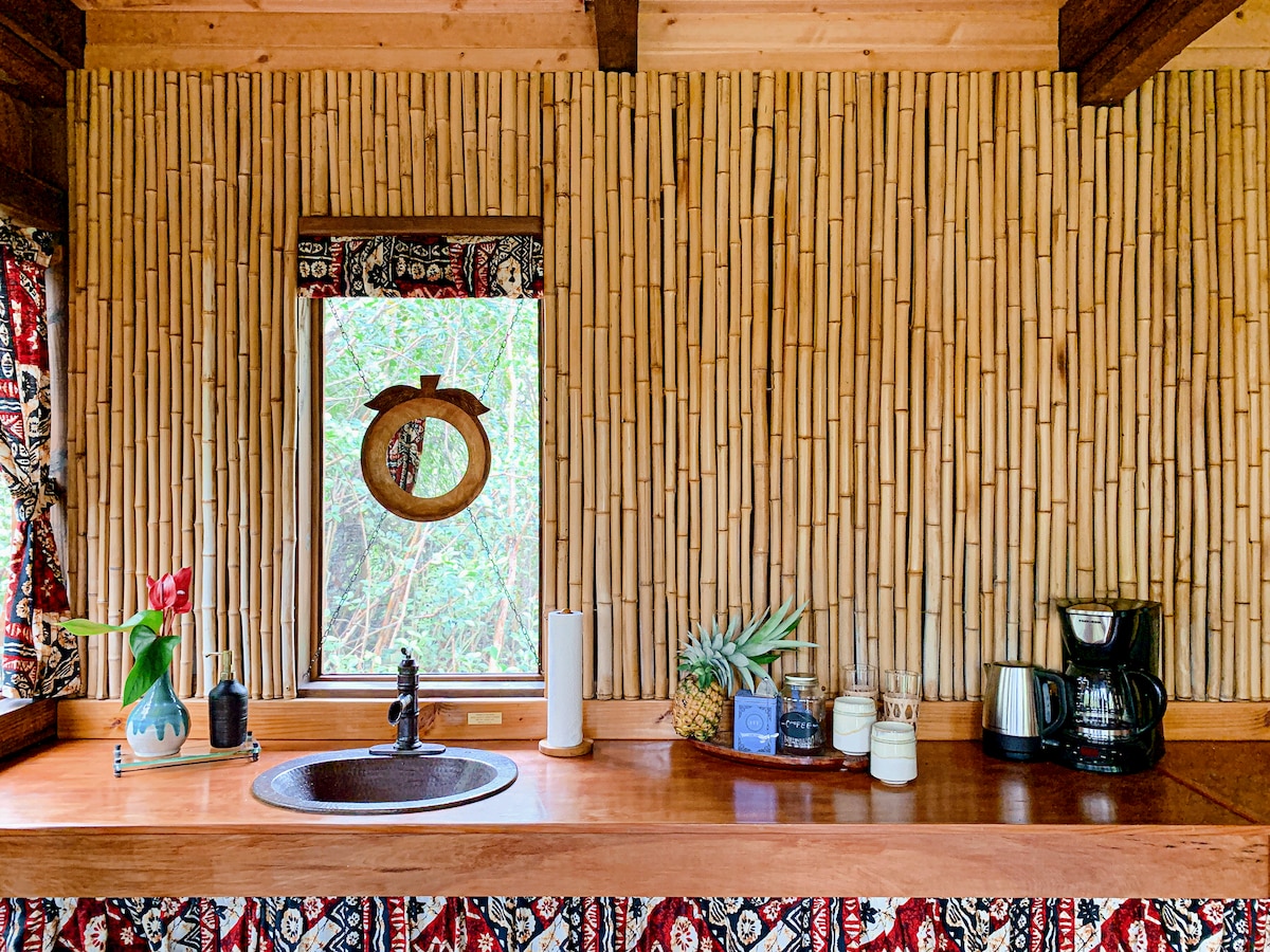 A kitchenette area features a bamboo wall, providing a natural aesthetic. A window offers views of the surrounding greenery, while a wooden countertop displays a coffee maker, a decorative pineapple, and a floral arrangement, contributing to a welcoming and functional space.