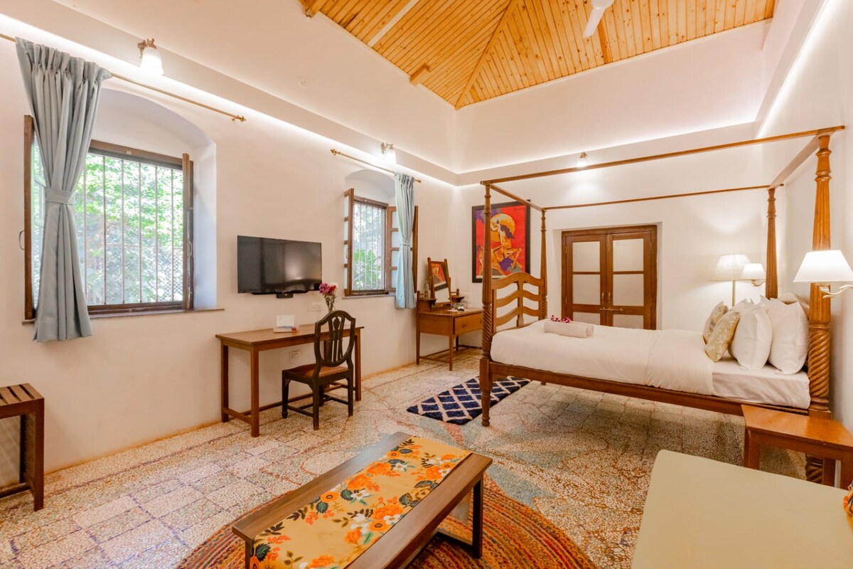 A spacious bedroom features a high wooden ceiling and large windows with light blue drapes. A four-poster bed is complemented by bedside lamps. A wooden desk and chair are positioned near a television, while a patterned rug adds warmth to the China mosaic flooring.