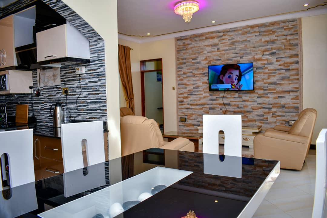The dining area features a sleek black table with white chairs, elegantly contrasting with the light-colored walls. A cozy seating area with a soft beige couch is visible, while a modern TV is mounted on a textured stone wall, enhancing the functional design.