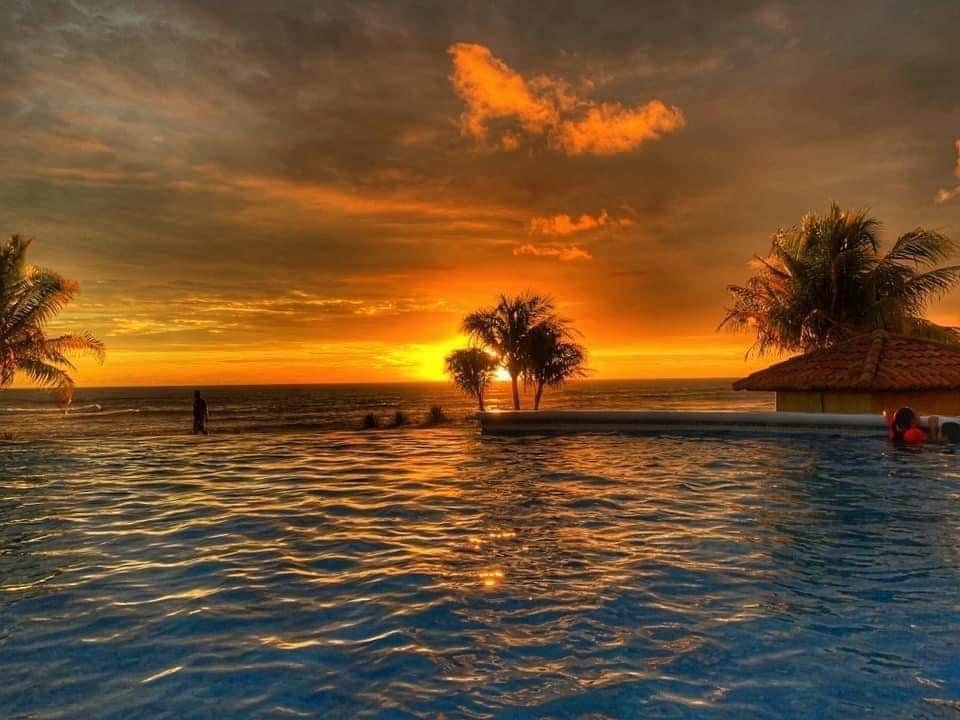 The infinity pool reflects a vibrant sunset, casting warm hues of orange and yellow across the water. Silhouetted palm trees frame the scene, providing a serene backdrop. Guests are seen enjoying the pool, with the horizon meeting the ocean in the distance.