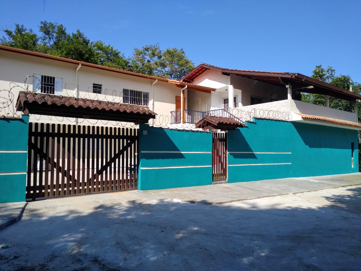 A welcoming exterior view features a turquoise wall with a wooden gate at the entrance. The two-story building showcases a terracotta roof and a balcony area. Green foliage is visible in the background, contributing to a peaceful atmosphere.