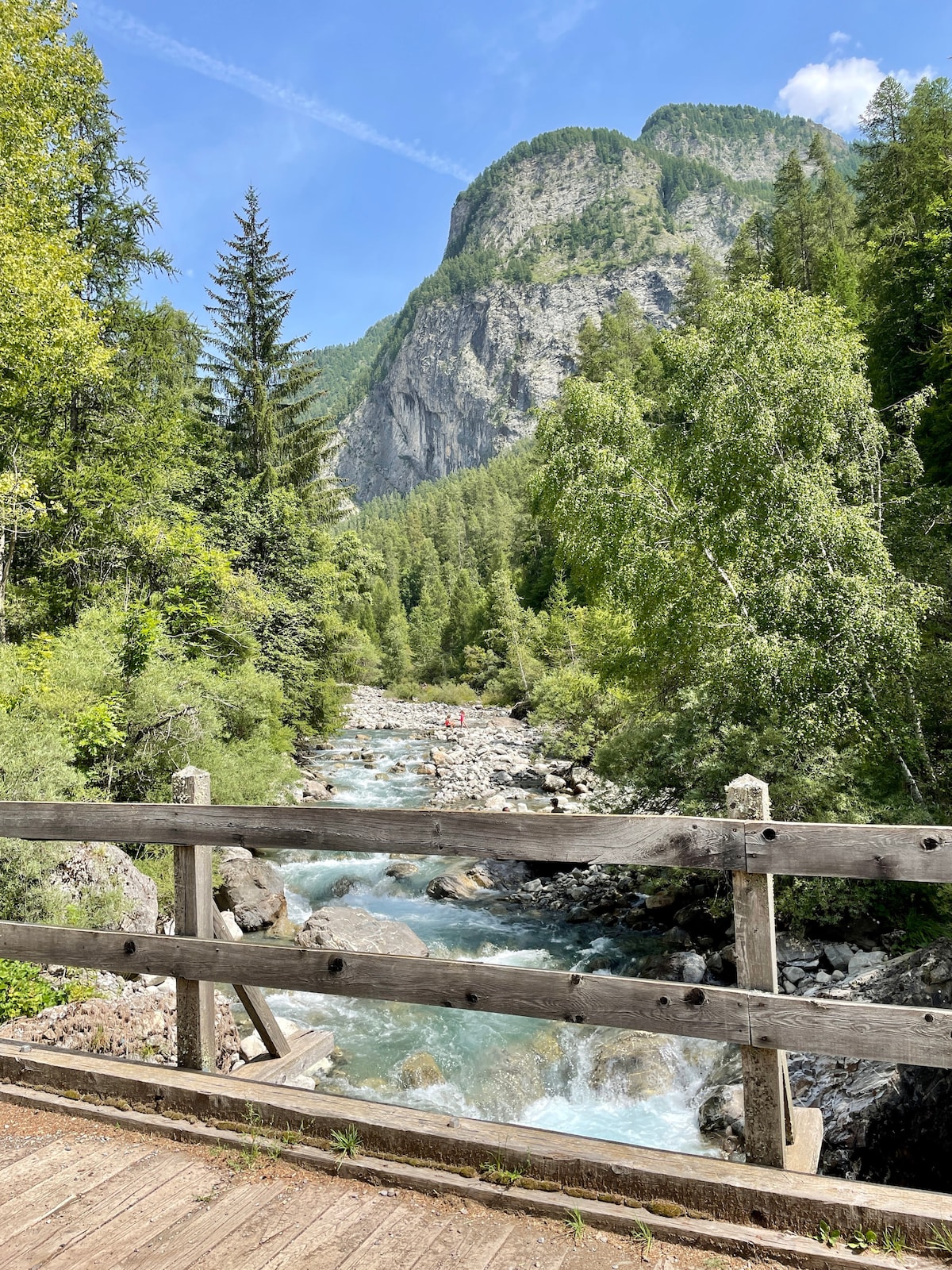 A wooden bridge spans a gently flowing river surrounded by lush greenery. Towering mountains rise dramatically in the background, partially obscured by trees. The vibrant blue water reflects the sky, and small figures can be seen enjoying the serene natural setting.