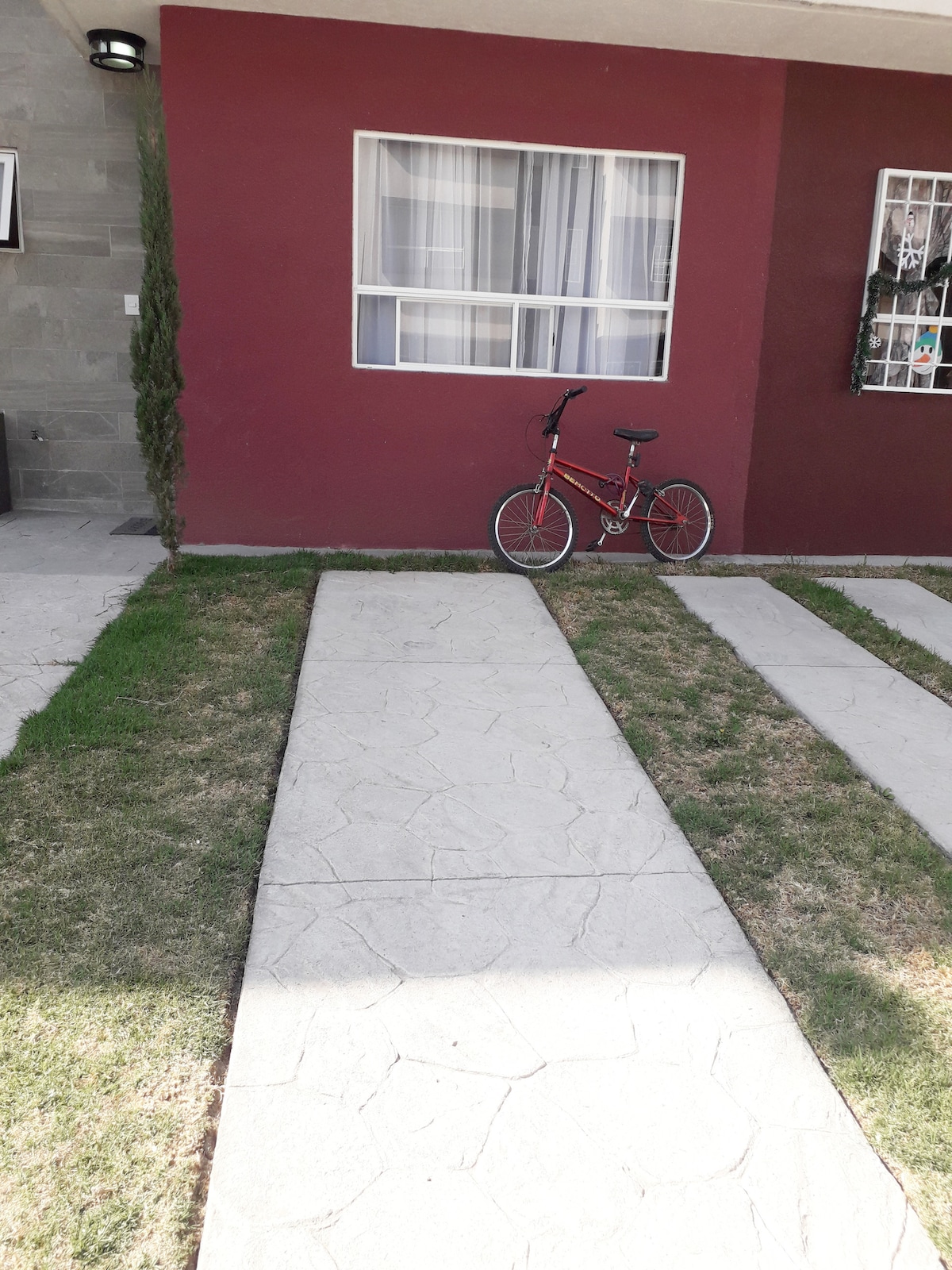 A well-maintained pathway of stone leads to the entrance of a home, bordered by neatly trimmed grass. A red bicycle is positioned beside a large window, which allows natural light to brighten the exterior.
