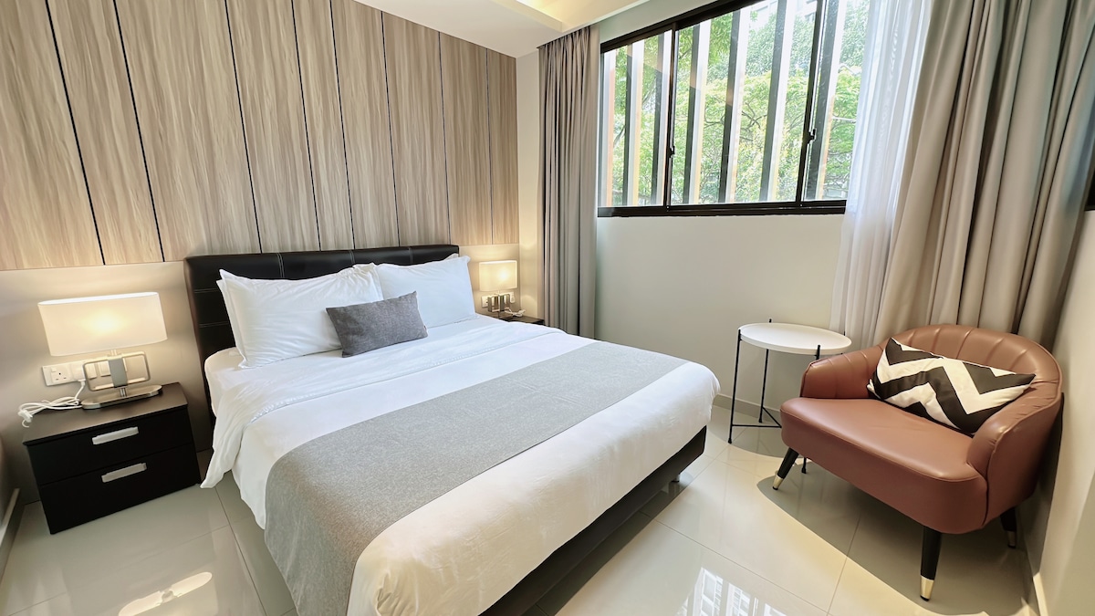 A cozy bedroom is highlighted by a spacious bed with light linens and a decorative pillow. A small round table and an armchair with a chevron pattern provide seating. Natural light filters through large windows bordered by soft curtains.