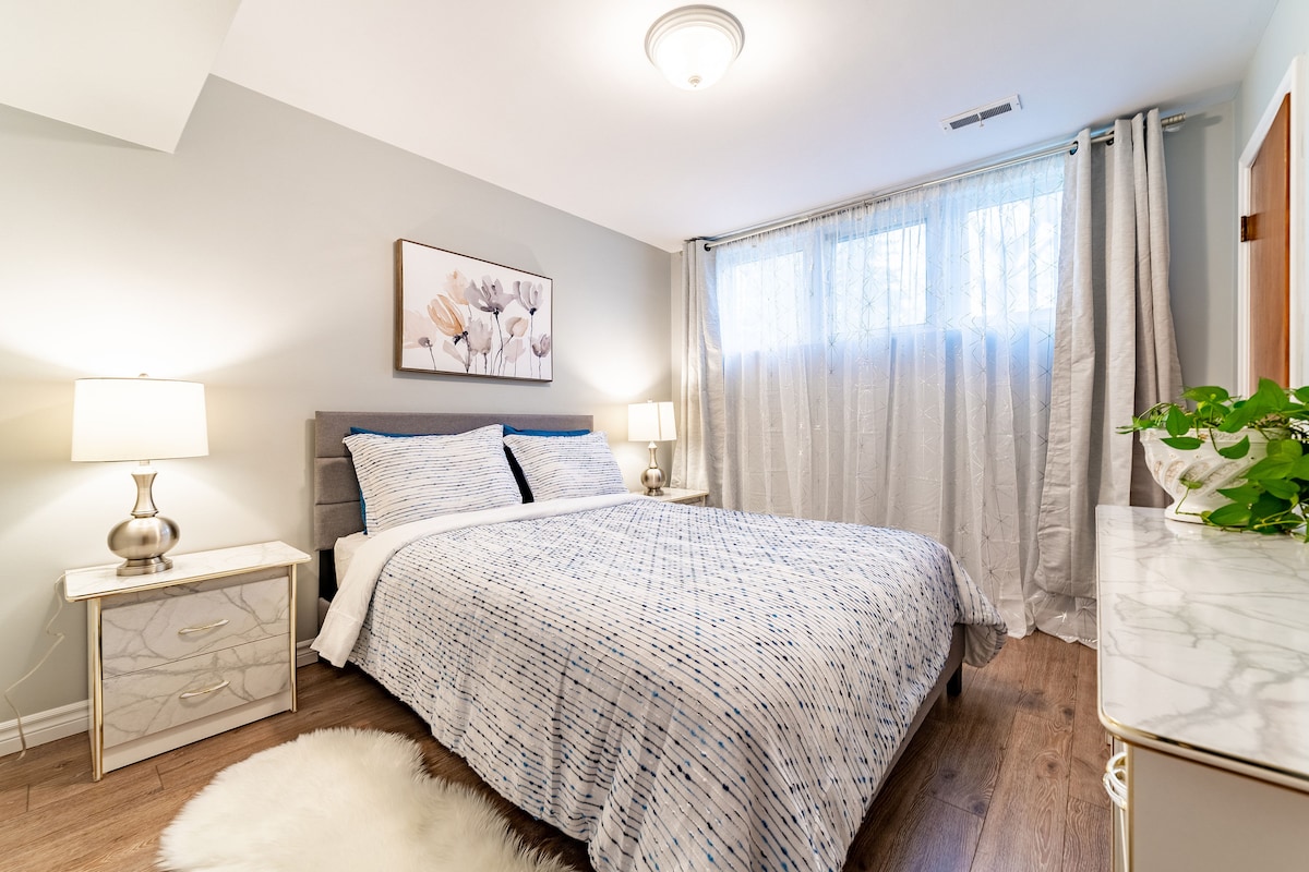 A serene bedroom features a spacious bed covered with a patterned blue and white quilt. Soft lighting is provided by bedside lamps on each side of the bed. Light filters through sheer curtains, while a small plant sits on the dresser against a neutral wall.