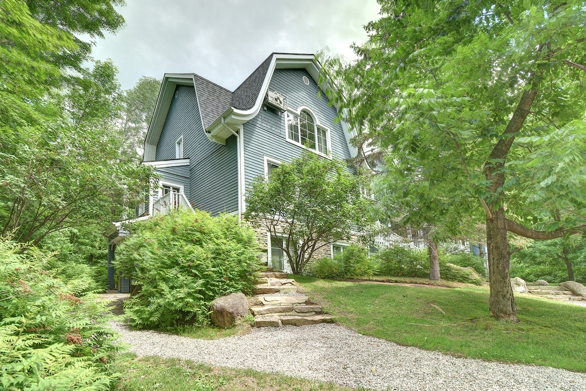 A charming blue exterior is framed by lush greenery and walking paths, offering a peaceful setting. Stairs and a gravel path lead up to the entrance, with a variety of trees providing natural shade around the property.