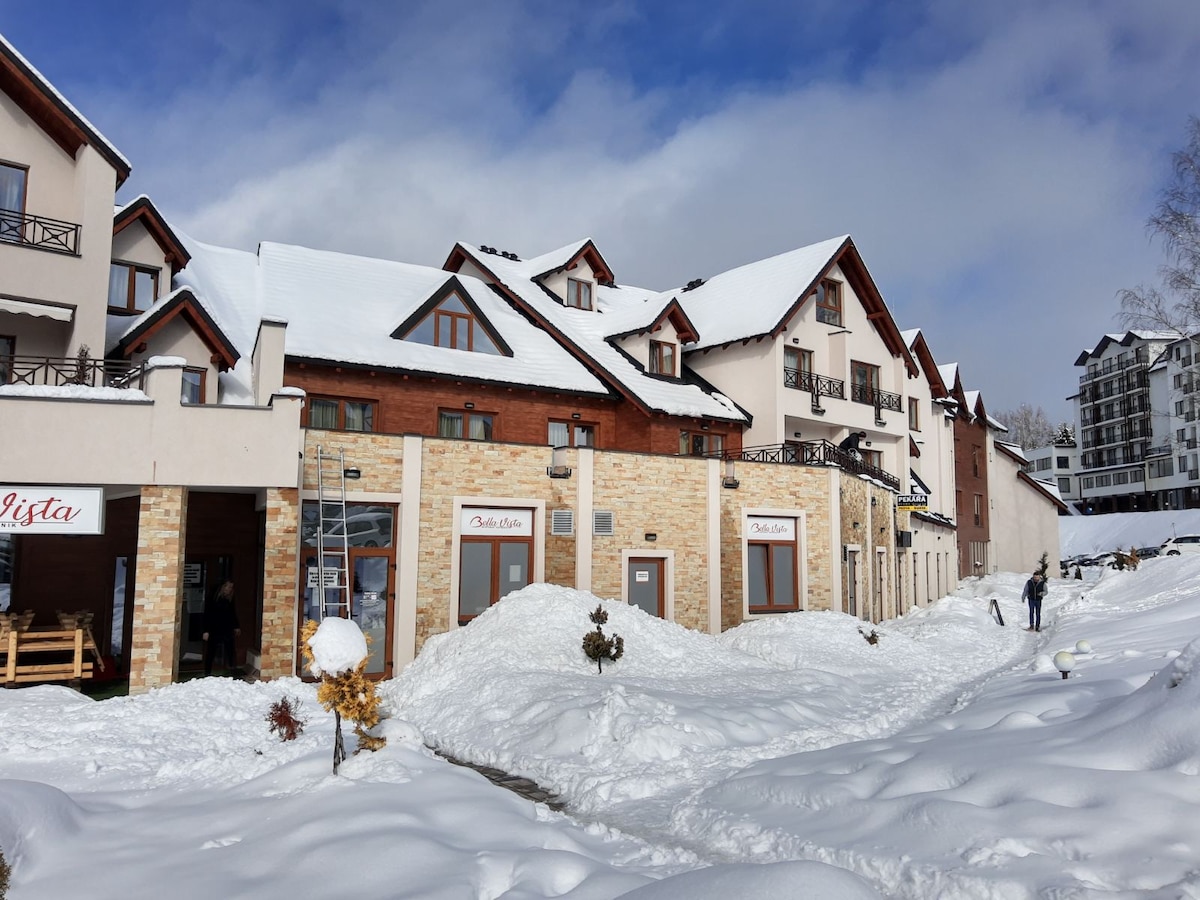 The image displays a charming resort complex, partially covered in snow, showcasing a blend of rustic and modern architectural styles. The entrance of a restaurant can be seen, along with snowy landscape features and mountains in the background, suggesting a serene winter atmosphere.